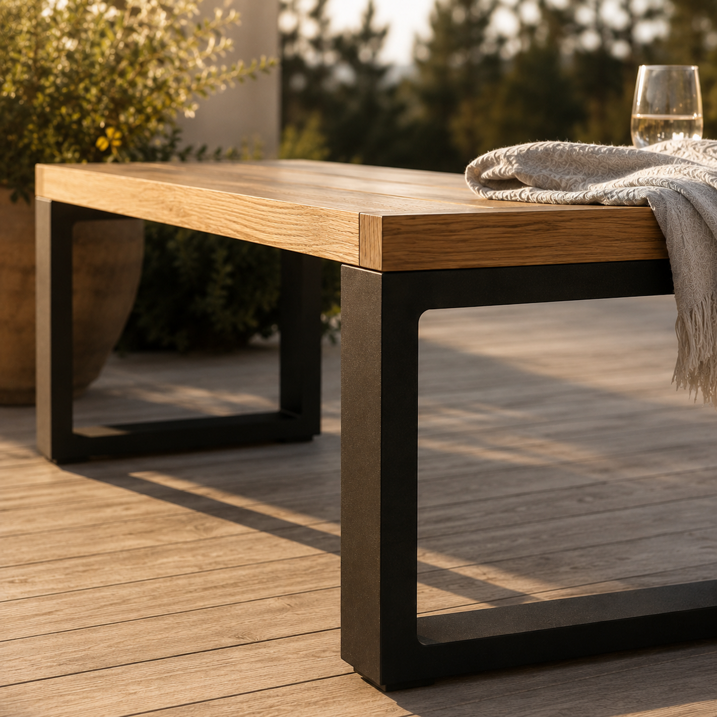 Detailaufnahme der modernen Sitzbank Urban aus massiver Eiche mit schwarzem Stahlgestell auf heller Holzterrasse im warmen Abendlicht. Sichtbar sind die präzise verarbeitete Holzplatte, klare Kanten und das minimalistische Kufengestell.