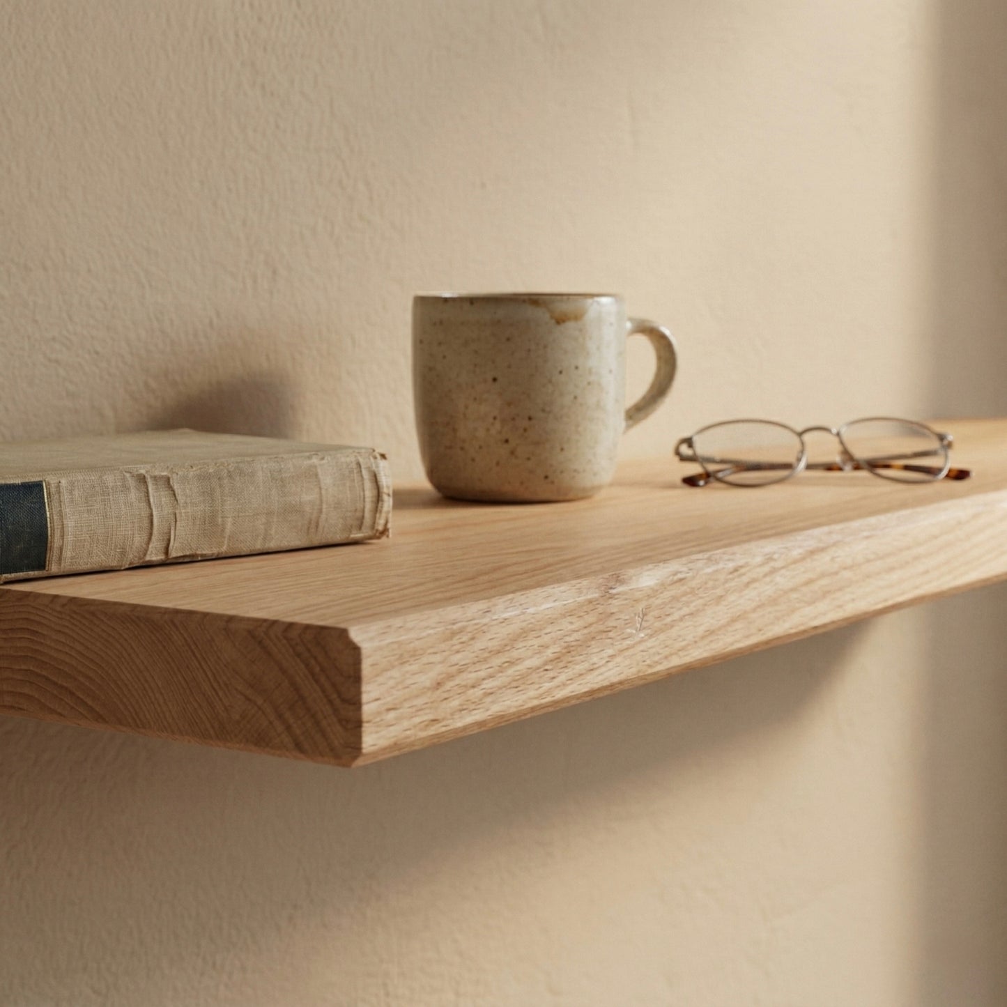 Detailaufnahme eines schwebenden Wandregals aus Buche furniert mit Buch, Keramiktasse und Brille in warmer, minimalistischer Umgebung