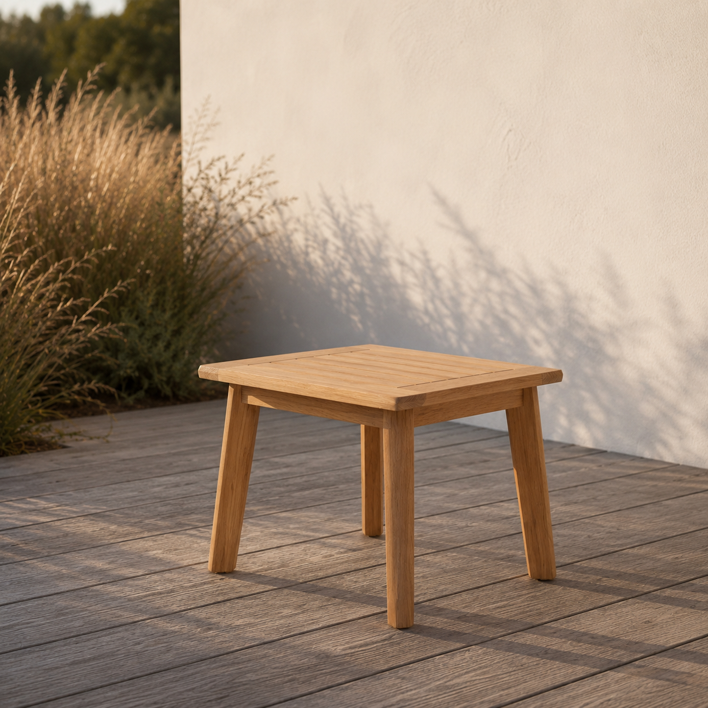 Quadratischer Sitzhocker Terra aus massiver Eiche auf moderner Holzterrasse im warmen Abendlicht. Minimalistische Outdoor-Szene mit heller Hauswand, natürlichen Gräsern und ruhiger mediterraner Atmosphäre.