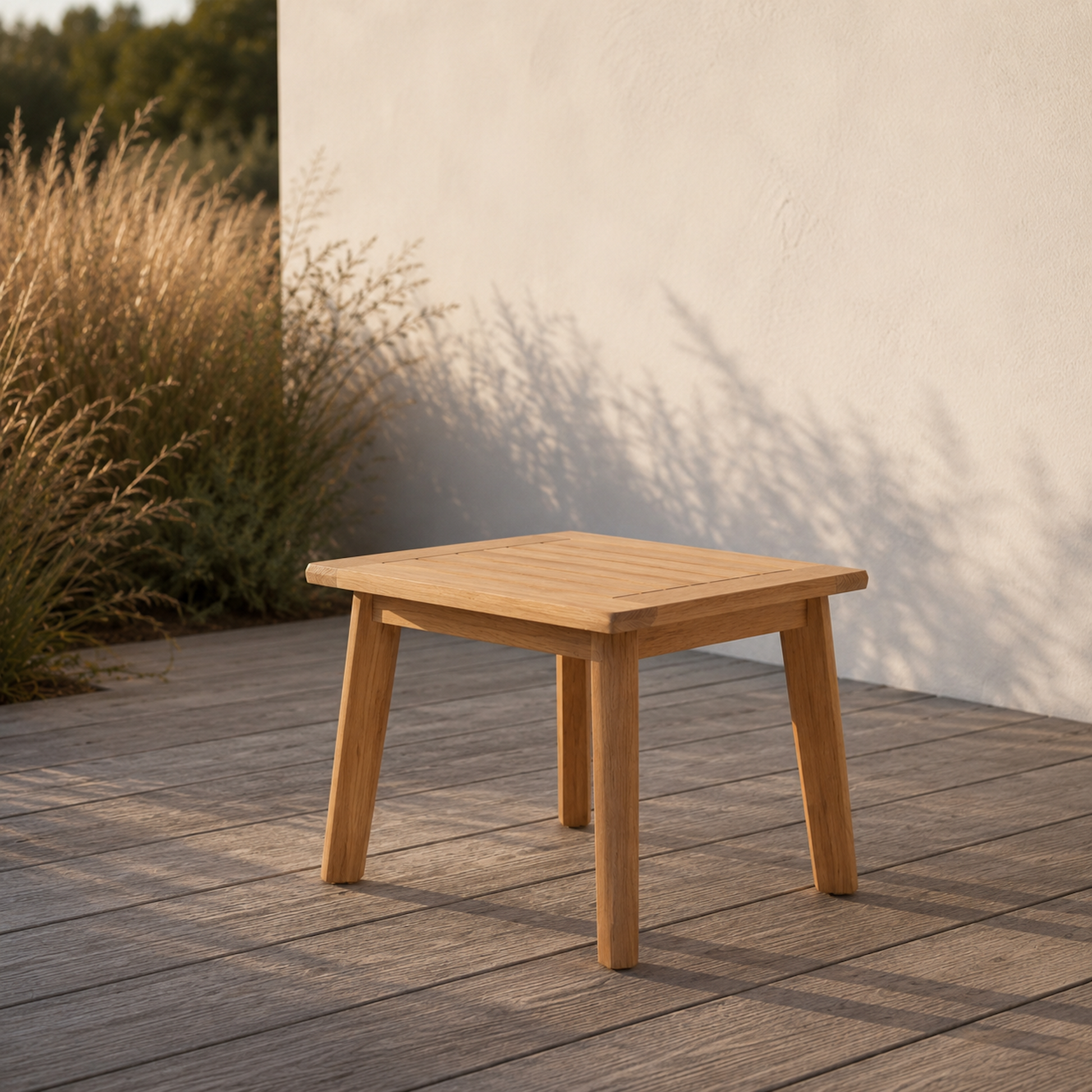 Quadratischer Sitzhocker Terra aus massiver Eiche auf moderner Holzterrasse im warmen Abendlicht. Minimalistische Outdoor-Szene mit heller Hauswand, natürlichen Gräsern und ruhiger mediterraner Atmosphäre.