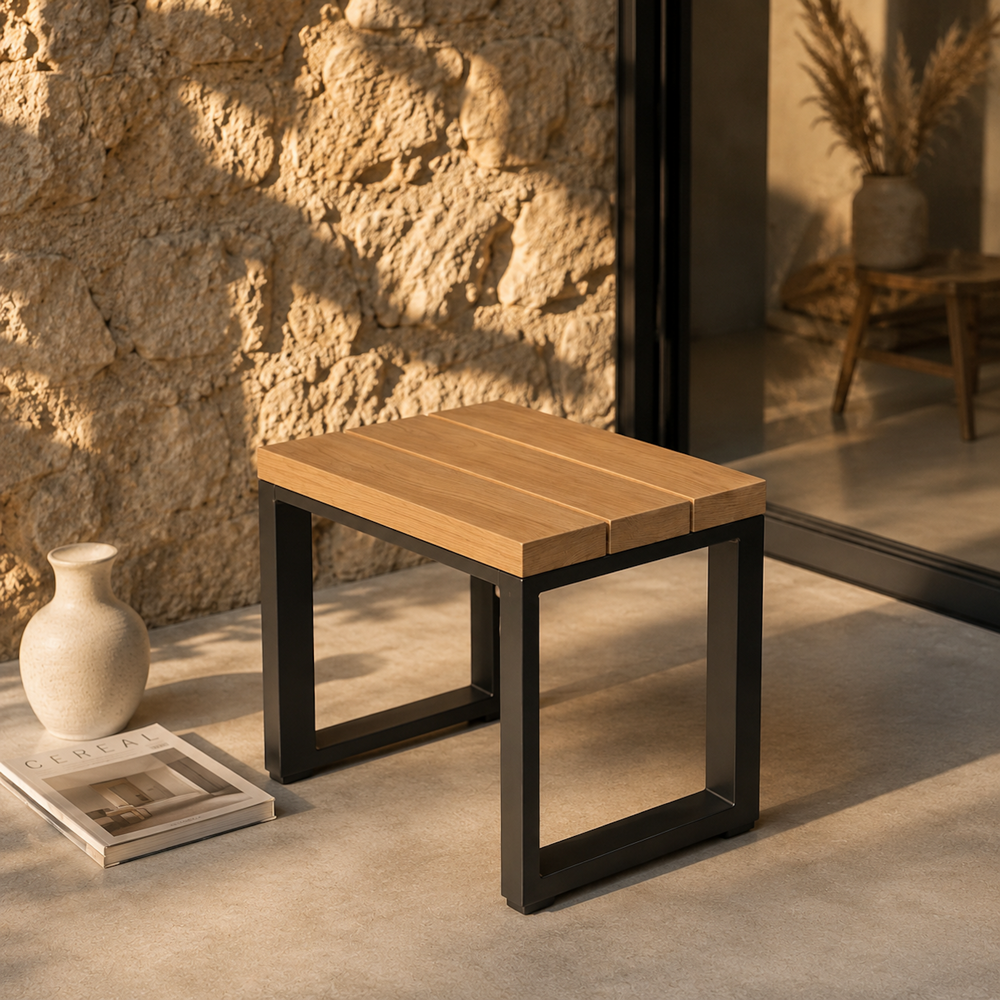 Moderner Sitzhocker Urban aus massiver Eiche mit schwarzem Stahlgestell auf einer minimalistischen Terrasse im warmen Abendlicht. Natürliche Steinwand, ruhige Outdoor-Atmosphäre und hochwertige Materialien im modernen Designstil.