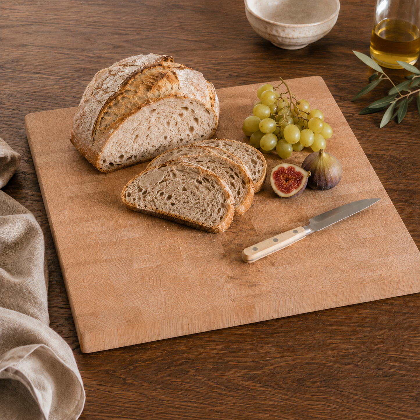 Ein großes Stirnholz-Schneidebrett aus Buche mit rustikalem Brot, grünen Trauben und Feigen auf dunklem Holztisch. Warmes Licht, hochwertiges Küchenmesser und natürliche Food-Styling-Elemente sorgen für eine gemütliche Atmosphäre.