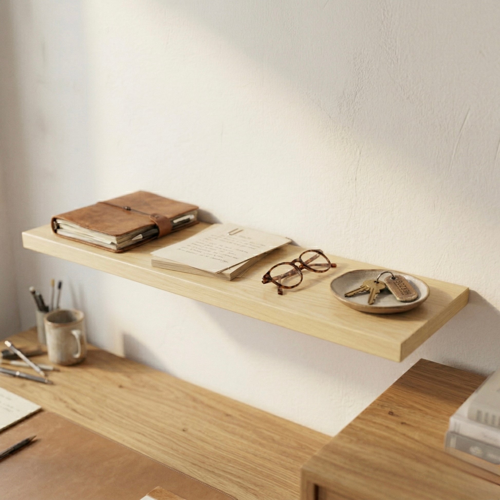 Schwebendes Wandregal aus Birke furniert über einem Schreibtisch mit Notizbuch, Brille und Schlüssel in einem warmen, minimalistischen Arbeitsbereich